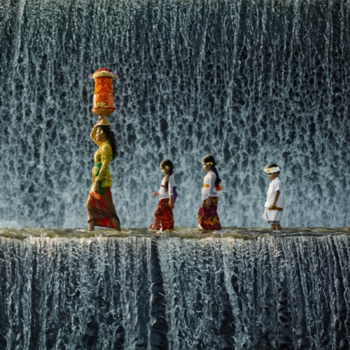 A woman and three children in traditional attire walk across a stone ledge against a cascading waterfall. The woman carries an ornate basket on her head.