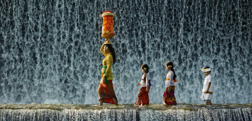 A woman and three children in traditional attire walk across a stone ledge against a cascading waterfall. The woman carries an ornate basket on her head.