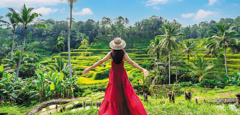 A woman in a red dress and straw hat stands with arms outstretched, facing lush green rice terraces and palm trees under a bright blue sky.