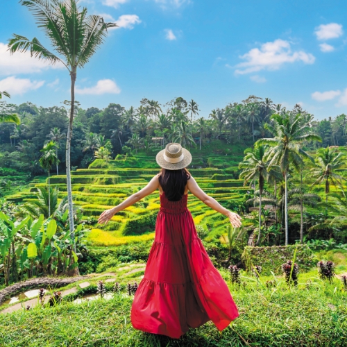 A woman in a red dress and straw hat stands with arms outstretched, facing lush green rice terraces and palm trees under a bright blue sky.