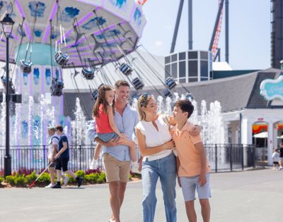 A family of four poses in front of a swing ride at an amusement park. The father holds a young girl, while the mother and a boy stand beside them. Fountains and other park visitors are visible in the background.