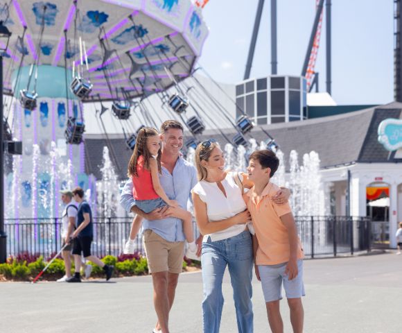 A family of four poses in front of a swing ride at an amusement park. The father holds a young girl, while the mother and a boy stand beside them. Fountains and other park visitors are visible in the background.
