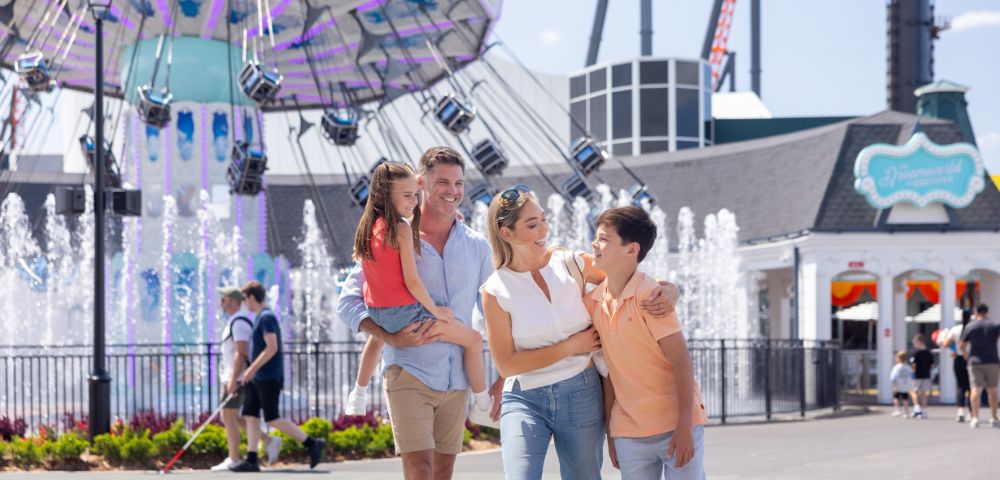 A family of four poses in front of a swing ride at an amusement park. The father holds a young girl, while the mother and a boy stand beside them. Fountains and other park visitors are visible in the background.