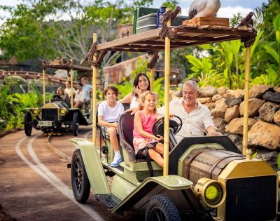Two vintage-style safari vehicles with open tops and wooden racks drive along a track in a lush, tropical environment. The front vehicle carries four passengers, and the rear one carries three. The scene is surrounded by greenery and rocks, with passengers' faces blurred for privacy.