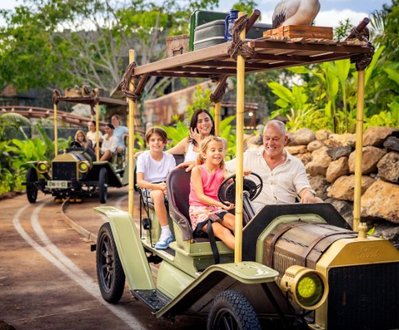 Two vintage-style safari vehicles with open tops and wooden racks drive along a track in a lush, tropical environment. The front vehicle carries four passengers, and the rear one carries three. The scene is surrounded by greenery and rocks, with passengers' faces blurred for privacy.