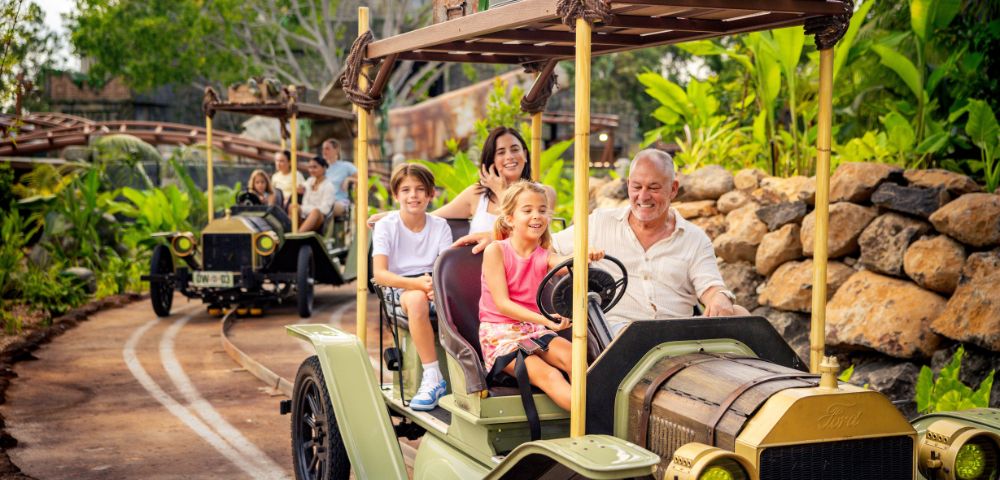Two vintage-style safari vehicles with open tops and wooden racks drive along a track in a lush, tropical environment. The front vehicle carries four passengers, and the rear one carries three. The scene is surrounded by greenery and rocks, with passengers' faces blurred for privacy.