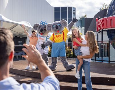 Two people in koala costumes wave on stage alongside two children and two adults, one of whom is taking a photo. A building with large windows and partial signage is visible in the background.