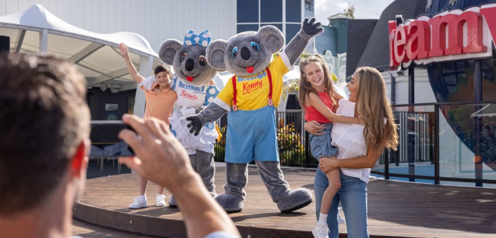 Two people in koala costumes wave on stage alongside two children and two adults, one of whom is taking a photo. A building with large windows and partial signage is visible in the background.