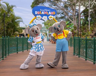 Two koala mascots, Kenny in a yellow shirt with red suspenders and blue shorts, and Belinda in a blue polka dot dress with a matching bow, stand on a wooden bridge at Dreamworld theme park. Above them is a sign that reads 'Kenny & Belinda's DREAMLAND' with colorful icons, surrounded by lush greenery and park attractions.
