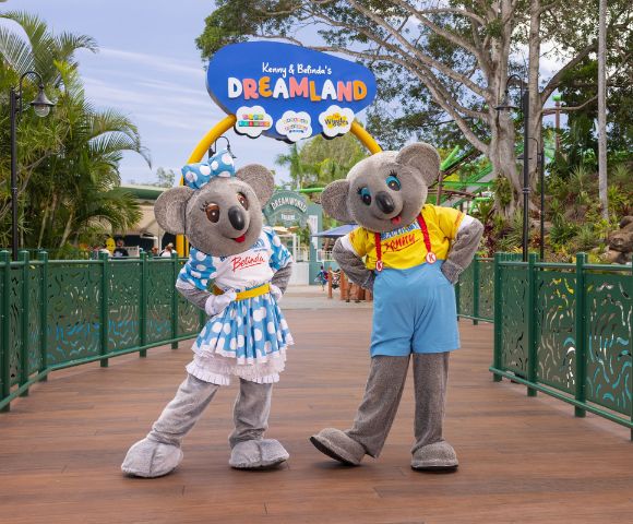 Two koala mascots, Kenny in a yellow shirt with red suspenders and blue shorts, and Belinda in a blue polka dot dress with a matching bow, stand on a wooden bridge at Dreamworld theme park. Above them is a sign that reads 'Kenny & Belinda's DREAMLAND' with colorful icons, surrounded by lush greenery and park attractions.