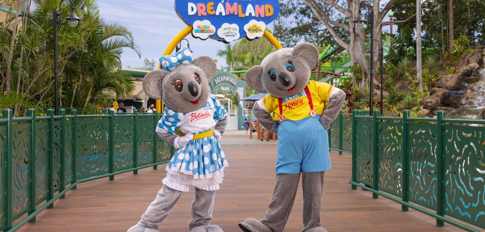 Two koala mascots, Kenny in a yellow shirt with red suspenders and blue shorts, and Belinda in a blue polka dot dress with a matching bow, stand on a wooden bridge at Dreamworld theme park. Above them is a sign that reads 'Kenny & Belinda's DREAMLAND' with colorful icons, surrounded by lush greenery and park attractions.