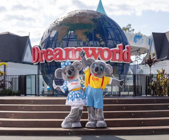 Two koala mascots, one named Belinda wearing a blue polka dot dress and bow, and the other named Kenny in a yellow shirt and blue shorts, stand in front of a large globe structure with a 'Dreamworld' sign in the background. Surrounding them are buildings with dark roofs and greenery.