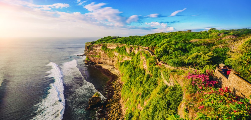 Scenic view of a lush green cliff overlooking the ocean at sunset. Waves crash against the rocks below. A path lined with colorful flowers runs along the edge.