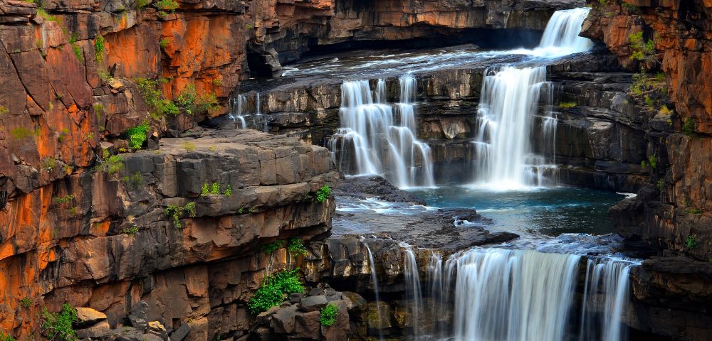 Multi-tiered waterfall cascading over rocky ledges surrounded by lush greenery.