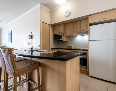 The image features a modern kitchen with a black countertop and wooden cabinets. The kitchen is equipped with a refrigerator on the right side, a microwave above the counter, and an oven below the counter. There is a sink located in the middle of the countertop, and a clock is mounted on the wall above it. In the kitchen, there are two chairs placed near the countertop, providing a comfortable seating area. A dining table can be seen in the background, with a chair positioned at the end of the table. The overall design of the kitchen is clean.