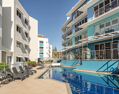 The image features a large swimming pool located in the middle of a city, surrounded by several buildings. The pool is filled with water, and there are multiple chairs placed around it, providing a comfortable spot for people to relax and enjoy the view. In addition to the chairs, there are a few dining tables scattered throughout the scene, suggesting that the area is also used for socializing and dining. A few people can be seen in the vicinity of the pool, possibly enjoying the sunny day or engaging in conversation.