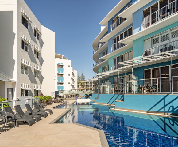The image features a large swimming pool located in the middle of a city, surrounded by several buildings. The pool is filled with water, and there are multiple chairs placed around it, providing a comfortable spot for people to relax and enjoy the view. In addition to the chairs, there are a few dining tables scattered throughout the scene, suggesting that the area is also used for socializing and dining. A few people can be seen in the vicinity of the pool, possibly enjoying the sunny day or engaging in conversation.