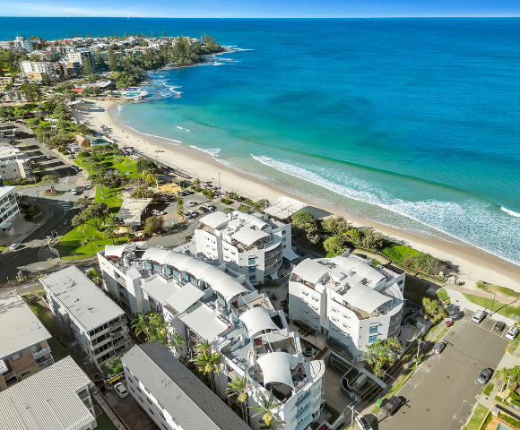 The image features a beautiful beach scene with a large body of water and a sandy beach. A row of white buildings, including apartments and houses, can be seen in the background, overlooking the beach. The buildings are situated on a hill, providing a stunning view of the ocean. In the foreground, there are several cars parked along the street, with a few boats visible in the water.