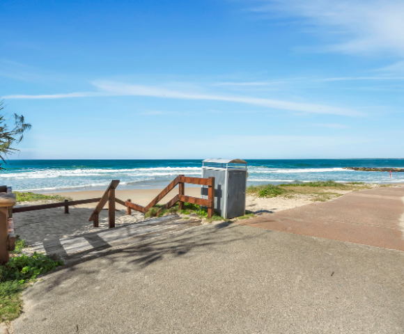The image features a picturesque beach scene with a wooden fence and a staircase leading down to the sandy shore. The stairs are positioned near a metal trash can, which is located close to the water's edge. The beach is surrounded by a peaceful ocean, with waves gently rolling in. In the background, there is a bench situated on the beach, providing a perfect spot for relaxation and enjoying the view. The overall atmosphere of the scene is serene and inviting, perfect for a day at the beach.