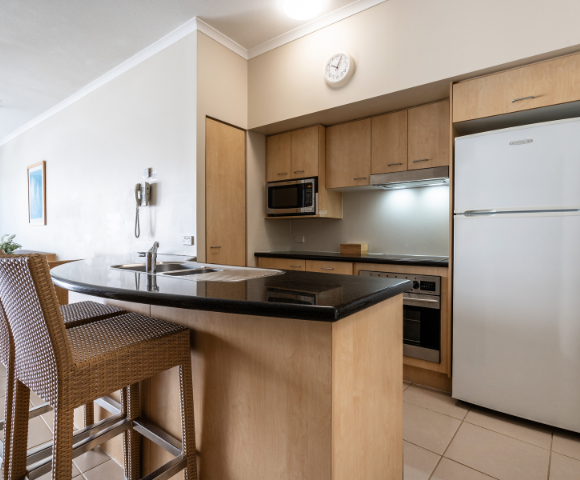 The image features a modern kitchen with a black countertop and wooden cabinets. The kitchen is equipped with a refrigerator on the right side, a microwave above the counter, and an oven below the counter. There is a sink located in the middle of the countertop, and a clock is mounted on the wall above it. In the kitchen, there are two chairs placed near the countertop, providing a comfortable seating area. A dining table can be seen in the background, with a chair positioned at the end of the table. The overall design of the kitchen is clean.
