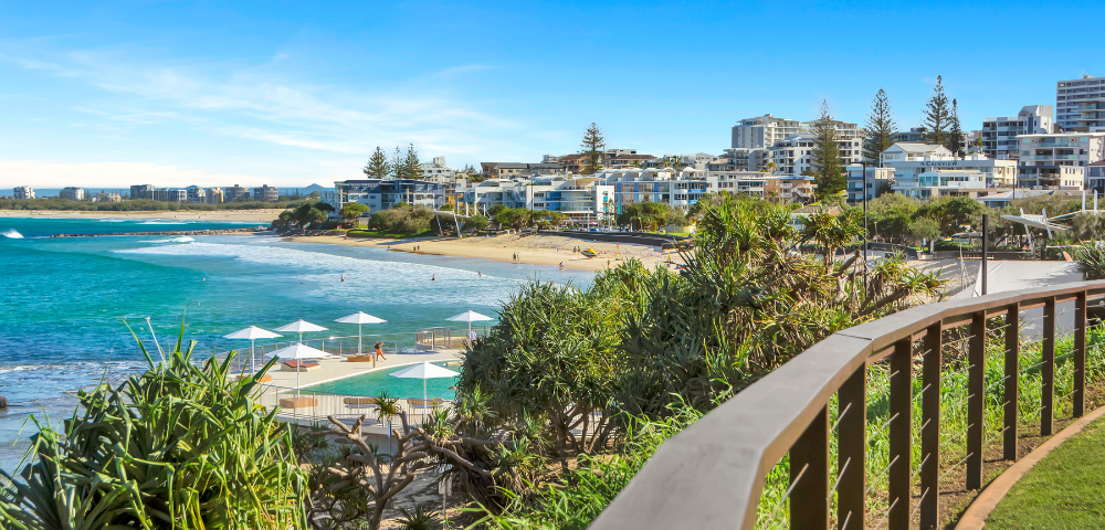 The image features a beautiful beach scene with a boardwalk overlooking the ocean. The boardwalk is lined with several umbrellas, providing shade for beachgoers. There are at least 13 umbrellas visible, scattered along the boardwalk at various distances from the water.
