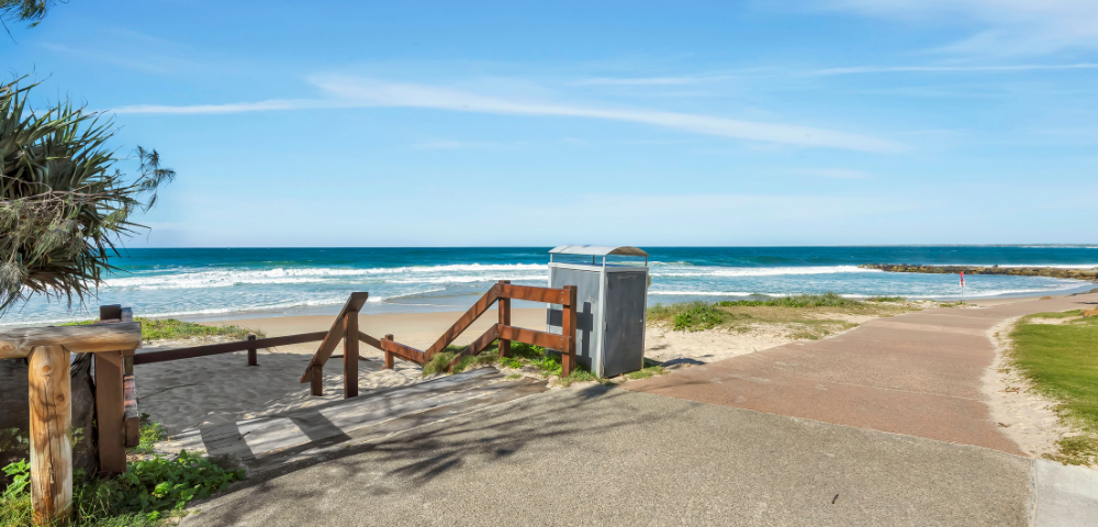 The image features a picturesque beach scene with a wooden fence and a staircase leading down to the sandy shore. The stairs are positioned near a metal trash can, which is located close to the water's edge. The beach is surrounded by a peaceful ocean, with waves gently rolling in. In the background, there is a bench situated on the beach, providing a perfect spot for relaxation and enjoying the view.