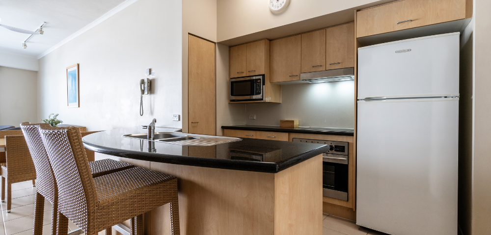 The image features a modern kitchen with a black countertop and wooden cabinets. The kitchen is equipped with a refrigerator on the right side, a microwave above the counter, and an oven below the counter. There is a sink located in the middle of the countertop, and a clock is mounted on the wall above it. In the kitchen, there are two chairs placed near the countertop, providing a comfortable seating area. A dining table can be seen in the background, with a chair positioned at the end of the table. The overall design of the kitchen is clean.