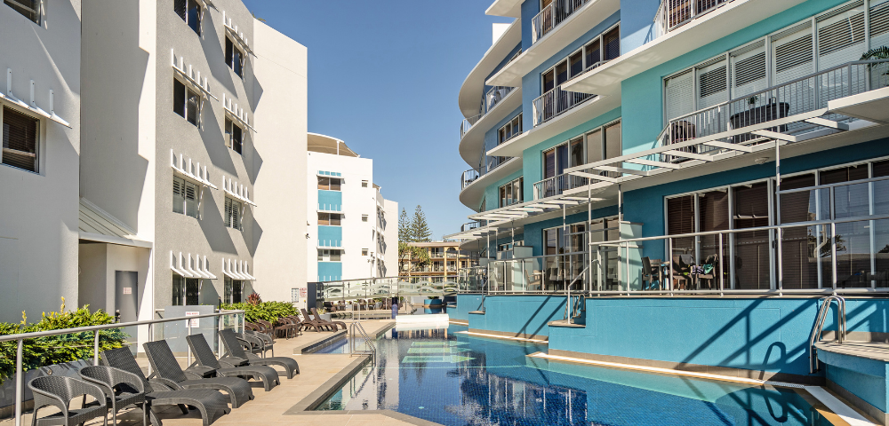 The image features a large swimming pool located in the middle of a city, surrounded by several buildings. The pool is filled with water, and there are multiple chairs placed around it, providing a comfortable spot for people to relax and enjoy the view. In addition to the chairs, there are a few dining tables scattered throughout the scene, suggesting that the area is also used for socializing and dining. A few people can be seen in the vicinity of the pool, possibly enjoying the sunny day or engaging in conversation.