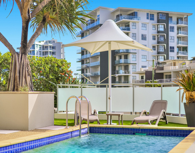 A sunny pool area featuring lounge chairs, a large umbrella, and tropical plants, set against modern apartment buildings.