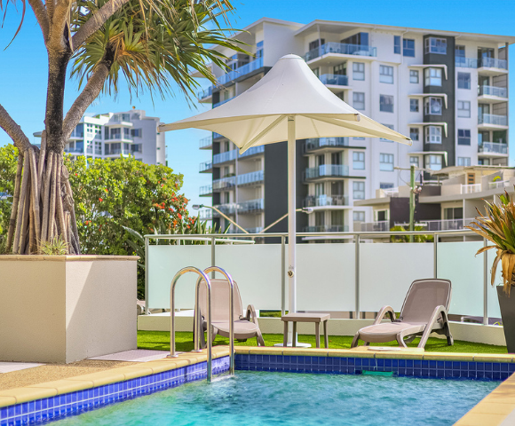 The image features a large swimming pool with a clear blue water surface, surrounded by a white fence. The pool is located in a backyard, with a building visible in the background. There are several chairs placed around the pool area, providing a comfortable spot for relaxation.