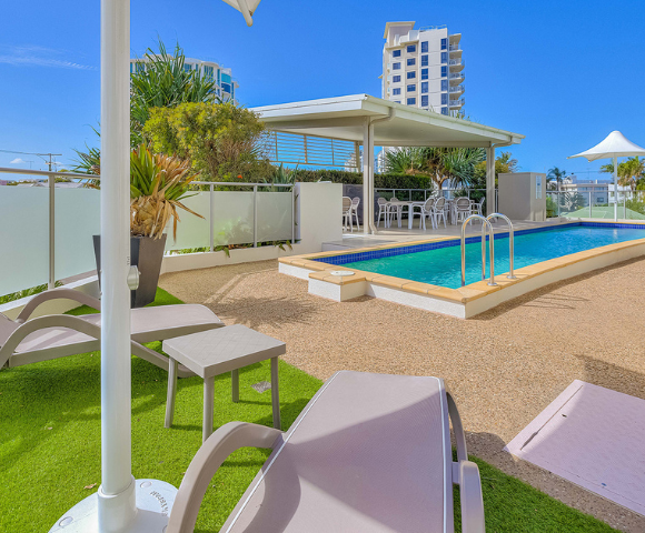 The image features a backyard with a large swimming pool, surrounded by several chairs and umbrellas. There are multiple chairs placed around the pool area, with some near the pool's edge and others further away. A few umbrellas are also present, providing shade for those relaxing by the pool.