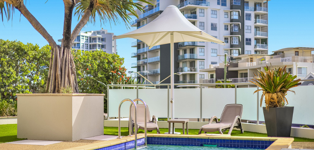 The image features a large swimming pool with a clear blue water surface, surrounded by a white fence. The pool is located in a backyard, with a building visible in the background. There are several chairs placed around the pool area, providing a comfortable spot for relaxation.