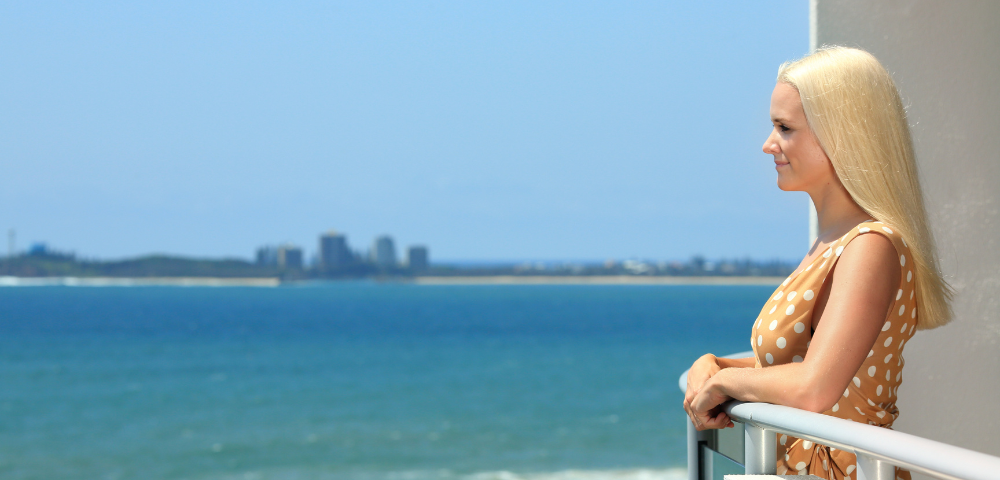 The image features a woman standing on a balcony overlooking a large body of water. She appears to be enjoying the view and the serene atmosphere. The balcony is located near a beach, providing a picturesque view of the ocean.