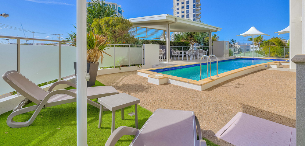 The image features a large swimming pool situated in front of a white apartment building. The pool is surrounded by several chairs, with some placed near the pool's edge and others scattered around the area. A dining table can also be seen close to the pool, providing a comfortable spot for outdoor dining or relaxation.