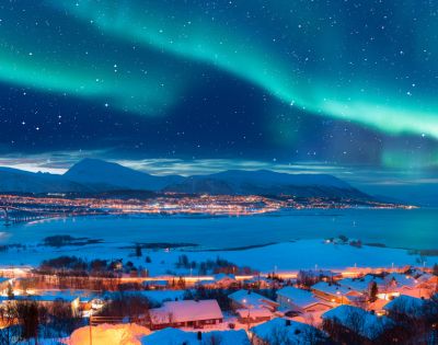 Vibrant auroras dance across a starry sky above a winter landscape, with a winding road and illuminated village by a fjord.