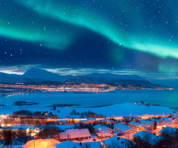Vibrant auroras dance across a starry sky above a winter landscape, with a winding road and illuminated village by a fjord.