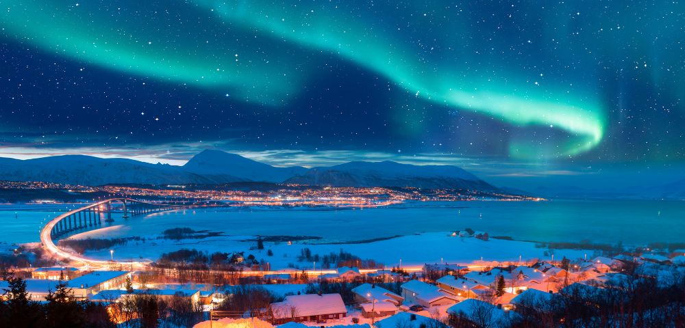 Vibrant auroras dance across a starry sky above a winter landscape, with a winding road and illuminated village by a fjord.