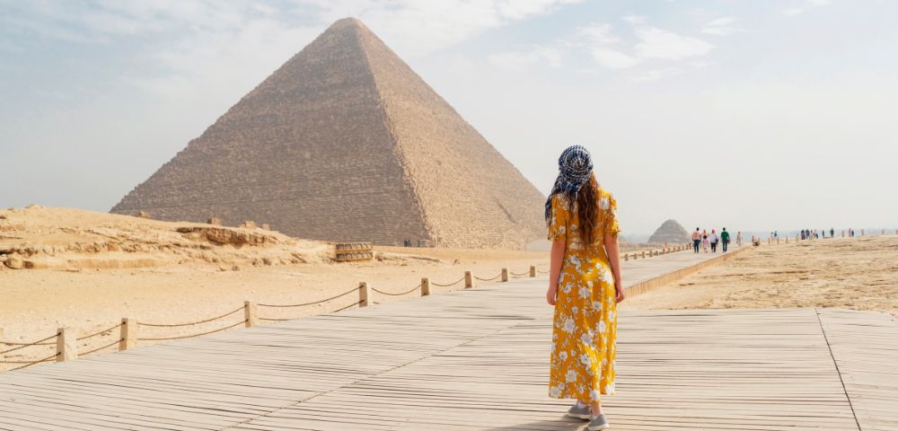 A woman in a yellow dress walks towards the Great Pyramid of Giza under a partly cloudy sky, conveying a sense of exploration and wonder.