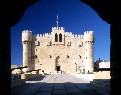 Sunlit medieval fortress with tall, rounded towers viewed from a shadowed archway. The sky is clear blue, enhancing the castle's grandeur and historical feel.