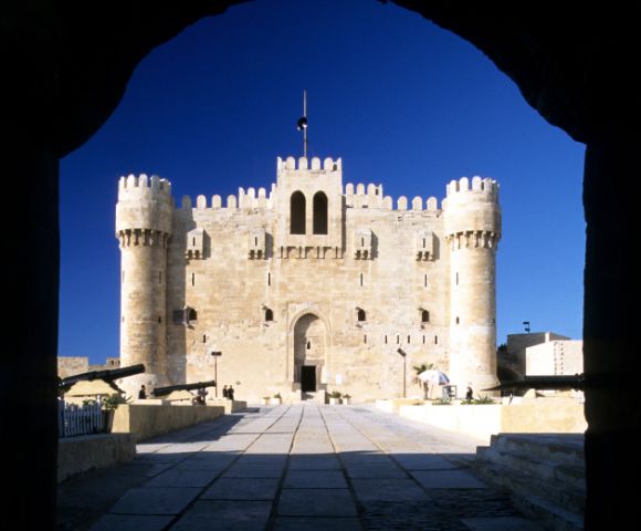 Sunlit medieval fortress with tall, rounded towers viewed from a shadowed archway. The sky is clear blue, enhancing the castle's grandeur and historical feel.