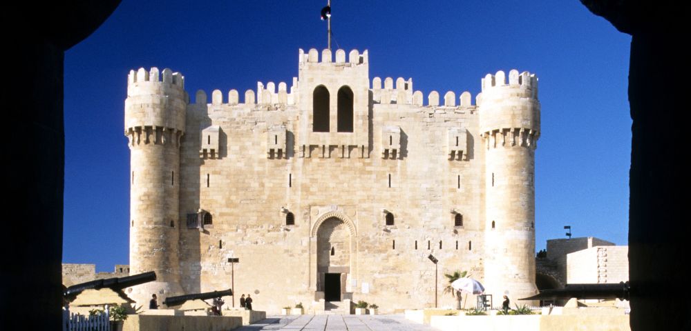 Sunlit medieval fortress with tall, rounded towers viewed from a shadowed archway. The sky is clear blue, enhancing the castle's grandeur and historical feel.