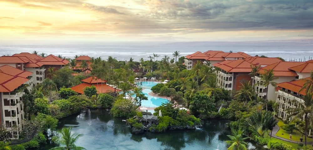 Tropical resort with red-roofed buildings, palm trees, and a serene pool by the ocean, bathed in warm sunset light.
