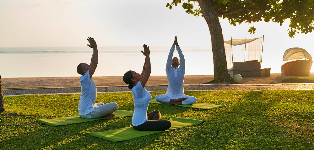 Three people practice yoga on green mats by the beach at sunrise, with a serene ocean backdrop and gentle waves lapping at the shore.