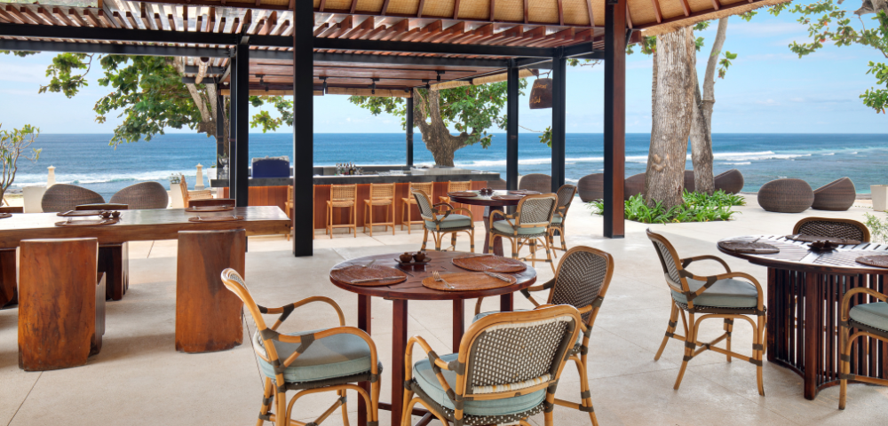 This image shows an open-air beachfront restaurant or café with a relaxed tropical atmosphere. The space has a thatched roof supported by dark wooden beams, giving it a natural and rustic feel. There are several round and rectangular wooden tables, paired with rattan-style chairs that have cushions for comfort. Toward the back, there is a bar counter with stools facing the ocean.