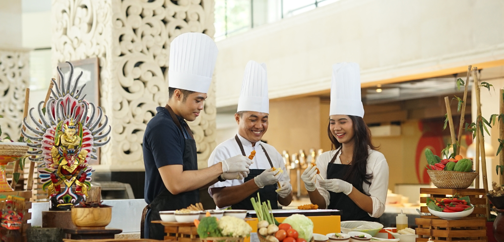 This image shows three people wearing chef hats and aprons, engaged in a cooking activity. Two of them are holding skewers with food, smiling as they prepare or decorate dishes together. The setting looks like a cooking class or a live culinary demonstration. The background includes a decorative carved wall panel, and the table in front of them is filled with fresh vegetables, herbs, and ingredients, suggesting a focus on traditional or cultural cuisine.