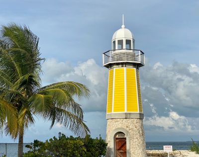 A yellow lighthouse with a stone base stands tall against a cloudy sky. A palm tree sways gently nearby, creating a serene coastal scene.