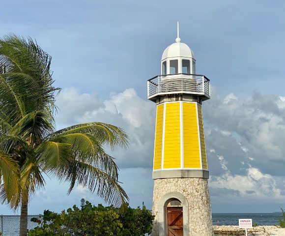 A yellow lighthouse with a stone base stands tall against a cloudy sky. A palm tree sways gently nearby, creating a serene coastal scene.