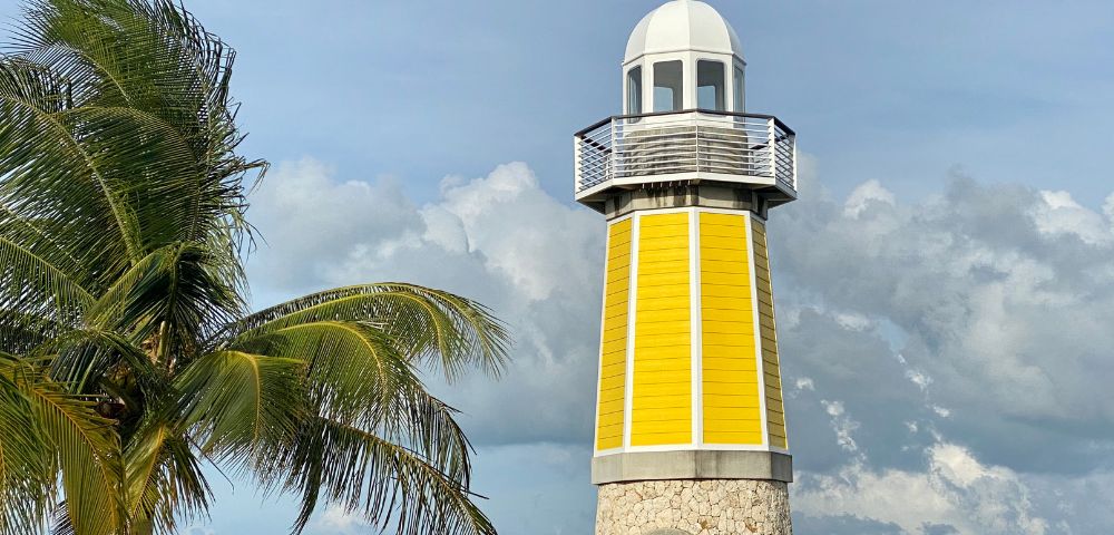 A yellow lighthouse with a stone base stands tall against a cloudy sky. A palm tree sways gently nearby, creating a serene coastal scene.