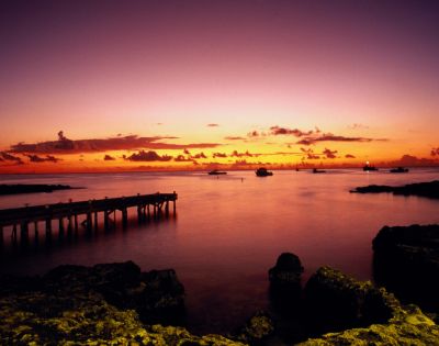 A tranquil sunset over a serene ocean, with a wooden pier to the left. Boats dot the horizon, and the sky transitions from orange to purple, creating a peaceful ambiance.