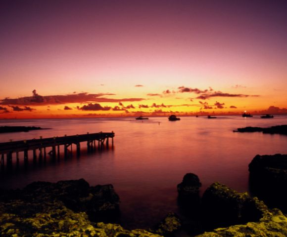 A tranquil sunset over a serene ocean, with a wooden pier to the left. Boats dot the horizon, and the sky transitions from orange to purple, creating a peaceful ambiance.